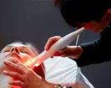 Une esthéticienne utilise un appareil Bloomea pour un soin du visage à la lumière rouge sur une cliente allongée.