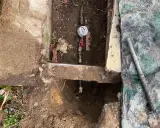 Réparation d'une fuite d'eau : un compteur et des tuyaux sont visibles dans une excavation, avec une bâche de protection à proximité.