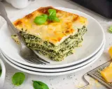 Ein Stück Spinatlasagne mit Käse überbacken, garniert mit Basilikum, auf einem Teller mit einer Gabel.