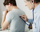 Arzt untersucht Patienten mit Stethoskop. Der Patient hustet, was auf Kurzatmigkeit hindeuten könnte.