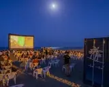 Noche de cine en el parque del cine Málaga. Público sentado en sillas en la playa, viendo una película en una pantalla gigante bajo la luna.