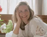 Glad kvinna med blont hår ler mot kameran, med handen mot kinden. En bukett blommor syns till vänster. Kanske ett foto från sofia wood blogg.