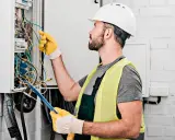 Elektryk w kasku i kamizelce odblaskowej pracuje przy skrzynce z przewodami. Zastanawia się, ile za punkt elektryczny.