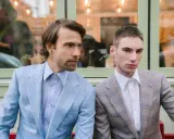 Two men in plaid suits with wide notch lapels sit side-by-side. The man on the left wears a light blue suit, the man on the right a tan suit.