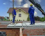 Dwaj pracownicy w niebieskich kombinezonach montują prefabrykowany strop. To kluczowy element budowy domu.