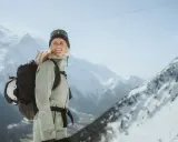 Kobieta w narciarskim stroju z plecakiem, do którego przymocowany jest kask, na tle gór.