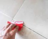 Mano aplica masilla con espátula roja para reparar una junta de baldosas dañada. Guía para cómo arreglar baldosas rotas.