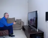 Starszy mężczyzna w okularach siedzi na kanapie i ogląda telewizję, trzymając pilota.