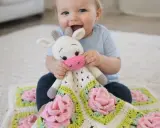 Bebé sonriente abraza una adorable manta de apego a crochet con forma de vaca y flores rosas.