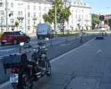Motocykl zaparkowany na chodniku, obok drogi z wyznaczonym pasem rowerowym. W tle budynki i ruch uliczny, w tym czerwony samochód i biały van.