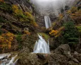 Cascada en un cañón rocoso, con árboles otoñales y niebla. El agua cae como un zafiro lagunazo, creando un espectáculo natural.