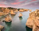 Felsformationen ragen aus dem ruhigen Wasser der **Lagoa, Portugal**, empor. Der Himmel ist in Rosa- und Orangetönen gefärbt, was auf einen wunderschönen Sonnenuntergang hindeutet.