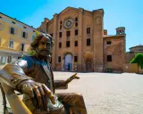 Statua bronzea in una piazza di una città dell'Emilia Romagna, con un'antica chiesa in mattoni sullo sfondo.