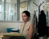 Una donna con un'espressione pensierosa siede in un ufficio disordinato, circondata da fascicoli. Sembra l'inizio di una storia nera.