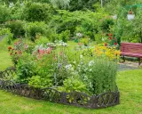 Un exuberante macizo de flores de verano, con rosas rojas, margaritas blancas y flores amarillas vibrantes, bordeado por una valla decorativa.