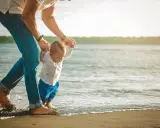 Padre ayuda a su bebé a dar sus primeros pasos en la orilla del mar, un recuerdo perfecto de Tarragona con niños.