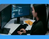 Jeune femme travaillant sur un ordinateur, avec deux écrans affichant du code. Sa position devant l'écran du bureau est concentrée.