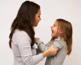 Mama śmieje się, łapiąc córkę za ręce. Dziecko radośnie szarpie się w nerwach, śmiejąc się głośno.
