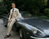 A man in a light-colored suit, perfect for mens summer cocktail attire, leans against a vintage car.