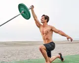 Mann führt eine Landmine Press-Übung am Strand aus, hebt eine Langhantel mit einem grünen Gewicht.