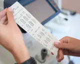 Dłonie trzymają wydruk z danymi optometrycznymi, gdzie widoczny jest numer paragonu.