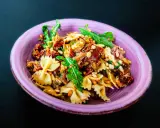 Ein köstlicher italienischer Nudelsalat mit Farfalle, sonnengetrockneten Tomaten, Pinienkernen und Rucola in einer lila Schüssel.