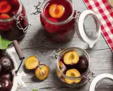 Frische Pflaumen werden für das Einkochen vorbereitet. Ganze und halbierte Früchte liegen neben Gläsern mit eingelegten Pflaumen.