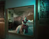 Elisa Miller sits on a bed, her reflection captured in a large mirror. The room features ornate wooden panels and a dramatic mountain mural.