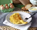 En gyllene omelett med ost, toppad med gräslök. Bredvid syns ägg, rostat bröd och mer ost. En perfekt start på dagen!