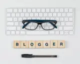 Okulary, klawiatura i drewniane klocki z napisem "BLOGGER" - idealne narzędzia do pisania bloga po angielsku.