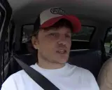 Mężczyzna w czapce z daszkiem i białej koszulce, siedzący w samochodzie. W tle widać drzewa i inne pojazdy. To może być początek jego zaorski wiek.