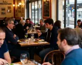 Des clients dînent dans une brasserie de gare, avec des assiettes de viande et des verres de vin sur la table.