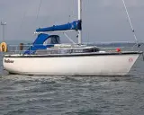 Voilier blanc Dufour 2800 à l'ancre, avec une bâche bleue sur la cabine. Un remorqueur orange est visible au loin.