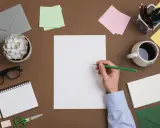 Student pisze list motywacyjny. Na biurku leżą koperty, notatnik, okulary, kubek kawy i przybory do pisania.