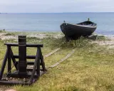 A dark wooden boat rests on a grassy shore next to a winch and wooden tracks leading to the sea.