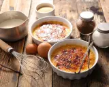 Dwa kremowe desery creme brulee z karmelizowanym cukrem, obok składniki: śmietanka, jajka, laski wanilii, cukier.