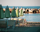 Ombrelloni verdi e lettini vuoti sulla spiaggia di una città riviera romagnola, con il mare calmo e una diga di sassi sullo sfondo.