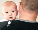 En söt bebis med stora ögon tittar fram över pappans axel. En liten vit fläck syns på pappans tröja, kanske från en rapning.