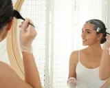 Frau hellt dunkle Haare auf, trägt Handschuhe und trägt Farbe mit einem Pinsel auf. Sie blickt lächelnd in den Spiegel.