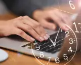 Dłonie piszą na klawiaturze laptopa, a nad nimi unosi się zegar. 0440 znaczenie czasu w pracy.