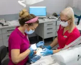 Dwie dentystki w maseczkach i rękawiczkach pracują przy pacjencie. Jedna z nich używa narzędzi, druga asystuje. Zabieg bez bólu dzięki lidokainie.