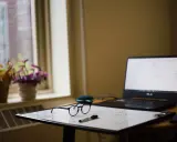 Okulary i pisak leżą na tablicy z zapiskami, obok laptopa. W tle okno z kwiatami. ibrus.