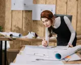 Architektka z rudymi włosami pochyla się nad planami na biurku, trzymając ołówek.