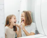 Mama i córka myją zęby, śmiejąc się. Dziecko nie chce myć zębów, ale mama przekonuje je zabawą.