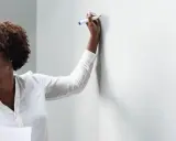 Frau mit Brille und Lockenwicklern hält einen Stift und bietet Nachhilfe an, während sie auf eine weiße Wand blickt.