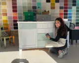 Mujer sonríe mientras trabaja en un mueble, aplicando pintura blanca. Aprende como pintar un mueble oscuro en blanco sin lijar.