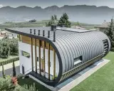 Nowoczesny dom z zaokrąglonym dachem z aluminium, które nie koroduje, otoczony zielenią i górami.