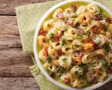 Kolorowa sałatka z tortellini, kawałkami kiełbasy i parmezanem, udekorowana natką pietruszki, podana na talerzu na zielonej serwetce w kropki.