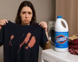 Smutna kobieta pokazuje ciemną koszulkę z pomarańczowymi plamami po wybielaczu. Obok butelka wybielacza Clorox i stos ubrań.