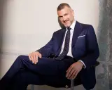 Uśmiechnięty mężczyzna w granatowym garniturze, z krawatem o klasycznej szerokości, siedzi w fotelu.